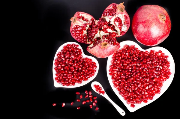 Two white heart shaped plates full of fresh ripe juicy pomegranate seeds, little spoon, whole fruit and ripe one on black background.