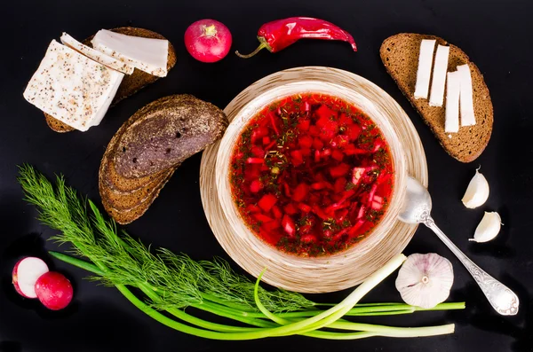 Beetroot soup in bowl with salted fresh lard, garlic, green onion, red pepper and rye bread on black background.