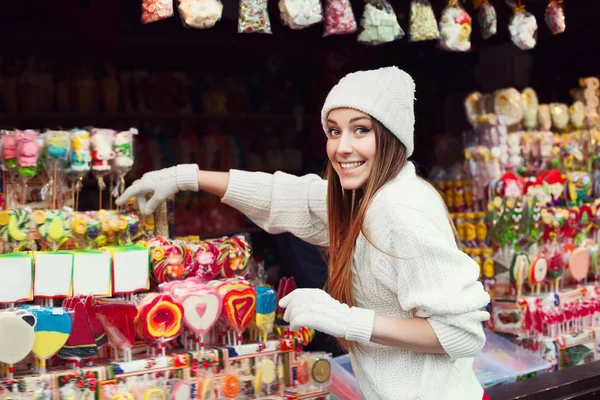 Street portrait of smiling beautiful young woman buying candy on the festive Christmas fair. Lady wearing classic stylish winter knitted clothes. Close up
