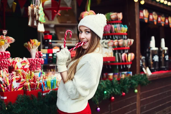 Street portrait of a smiling beautiful young woman biting candy cane and looking at camera. Lady wearing classic winter knitted clothes. Festive Christmas fair as background. Close up