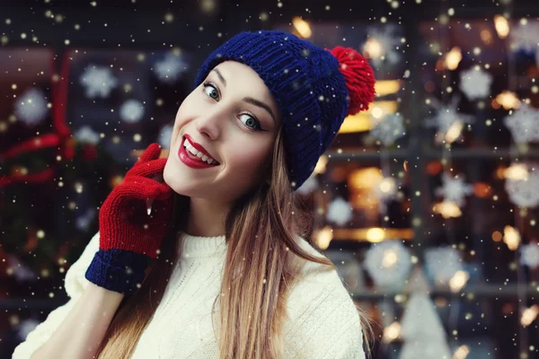 Street portrait of smiling beautiful young woman wearing classic winter knitted clothes. Model looking at camera. Festive garland lights. Magic snowfall effect. Close up. Toned