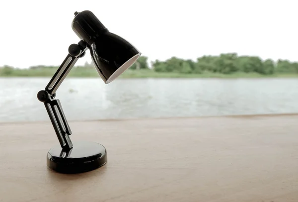 Small black lamp on wooden table with river and forest as background in low key tone. Lamp is compared to a man in gloomy and sad feelings.