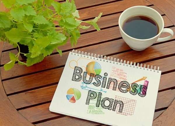 Notebook with text inside Business Plan on table with coffee and plant