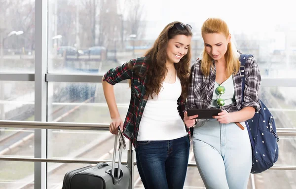 Two girls stand with suitcases at the airport and looking at the tablet. A trip with friends