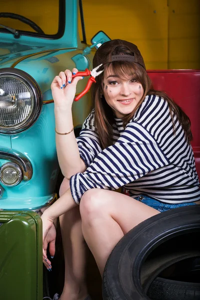 Young attractive woman in a baseball cap sits in the garage near the retro car with tools. Girl holding pliers