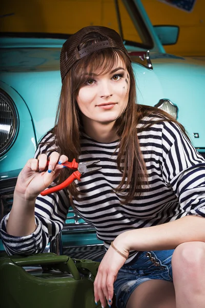 Young attractive woman in a baseball cap sits in the garage near the retro car with tools. Girl holding pliers