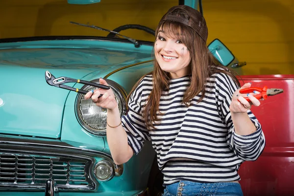 Cheerful woman sits in the garage near the retro car with tools. Girl holding pliers and an adjustable wrench
