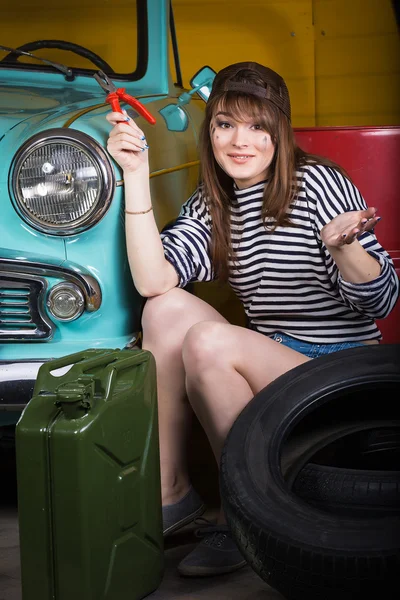 Young attractive woman in a baseball cap sits in the garage near the retro car with tools. Girl holding pliers