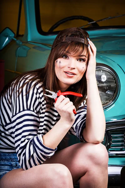Woman in a cap sits tired in the garage near the retro car with tools. Girl holding pliers and took hold of the head