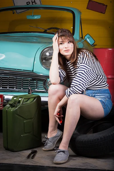 Woman in a cap sits tired in the garage near the retro car with tools. Girl holding pliers and took hold of the head