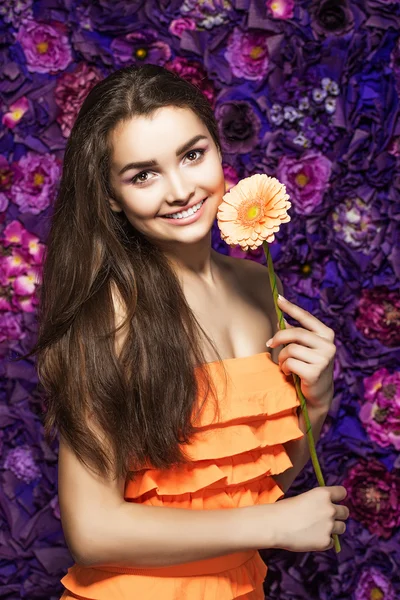 Brunette woman with a gentle make-up that looks at the camera while holding flower near the face on a floral background.