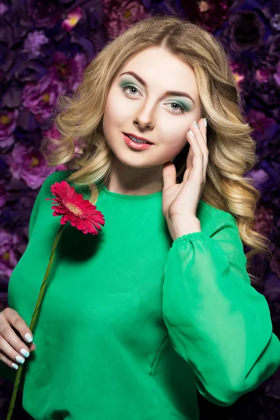 Blonde woman with a gentle make-up that looks at the camera while holding flower near the face on a floral background.