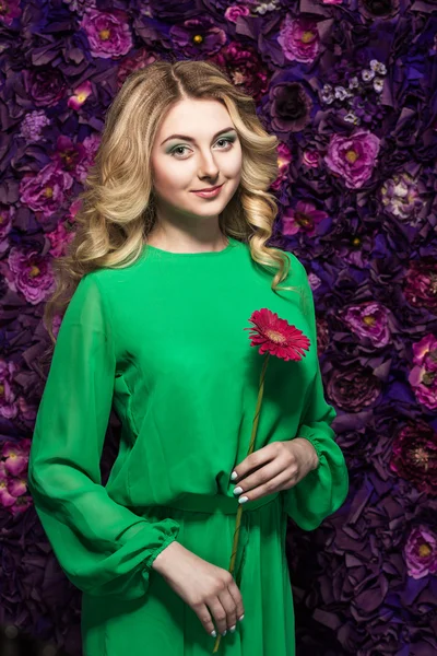 Blonde woman with a gentle make-up that looks at the camera while holding flower near the face on a floral background.