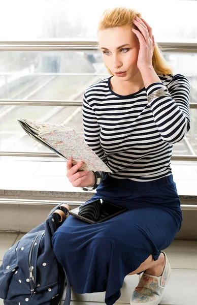 Woman in striped blouse sitting at the station or terminal and looking at the map and holding his head in surprise