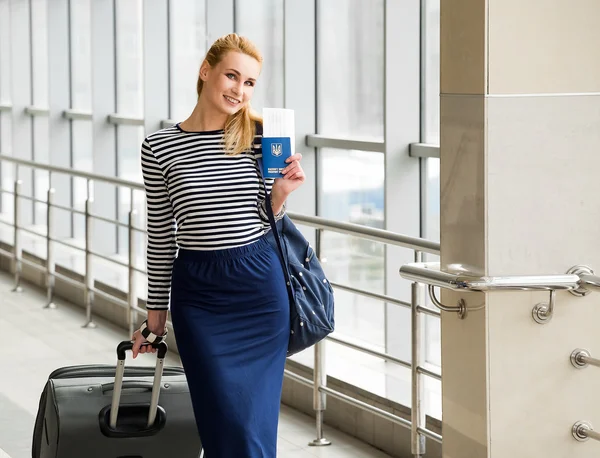 Tourist blonde girl in a striped blouse standing on the station with a large suitcase and passport with tickets
