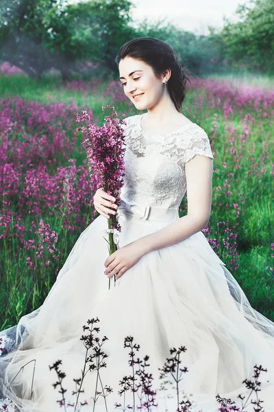 Beautiful bride in a flower field. The girl in a white dress with a bouquet in a summer field at sunset