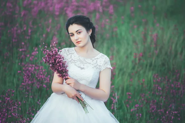 Beautiful bride in a flower field. The girl in a white dress with a bouquet in a summer field at sunset