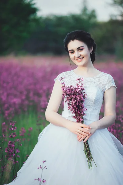 Beautiful bride in a flower field. The girl in a white dress with a bouquet in a summer field at sunset