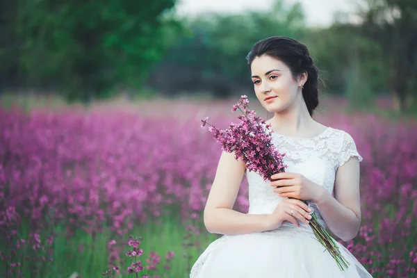 Beautiful bride in a flower field. The girl in a white dress with a bouquet in a summer field at sunset