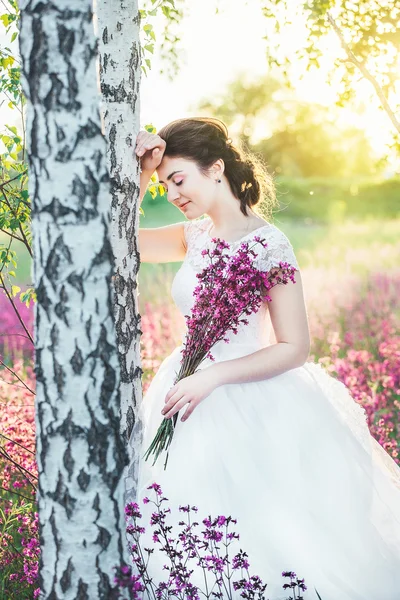 The girl in a white dress with a bouquet in a summer field at sunset stands near the birch