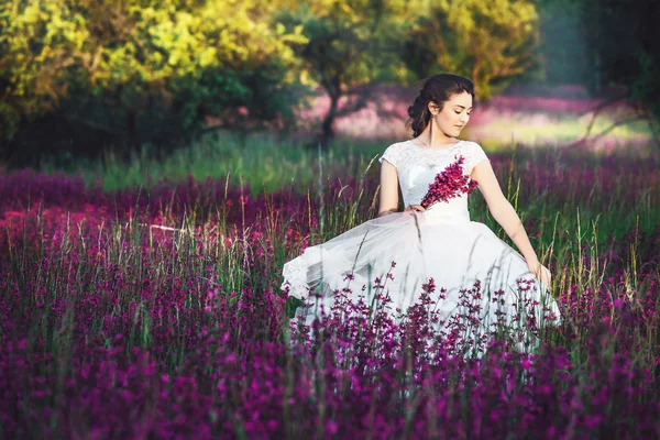 Beautiful bride in a flower field. The girl in a white dress with a bouquet in a summer field at sunset