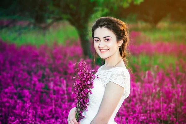 Beautiful bride in a flower field. The girl in a white dress with a bouquet in a summer field at sunset