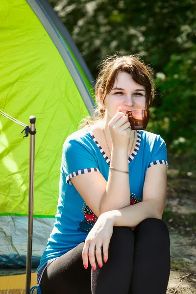 Young brunette woman in blue blouse sitting in a forest near the tent.tourist resting on a halt near a fire with smoke.