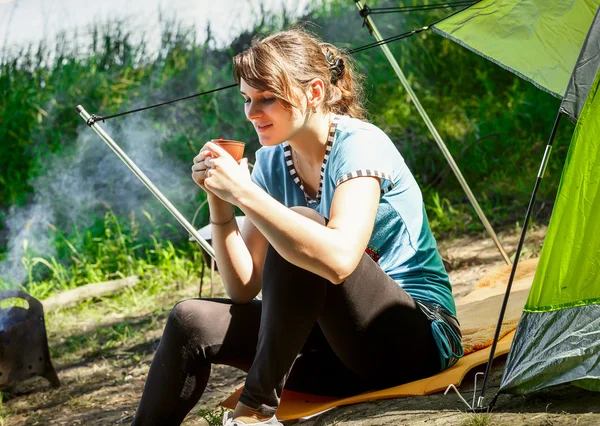 Young brunette woman in blue blouse sitting in a forest near the tent.tourist resting on a halt near a fire with smoke.