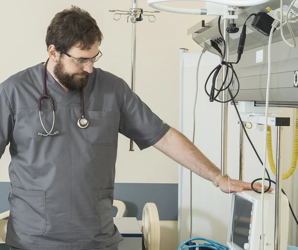 Bearded doctor wearing glasses and a gray robe works with hospital equipment