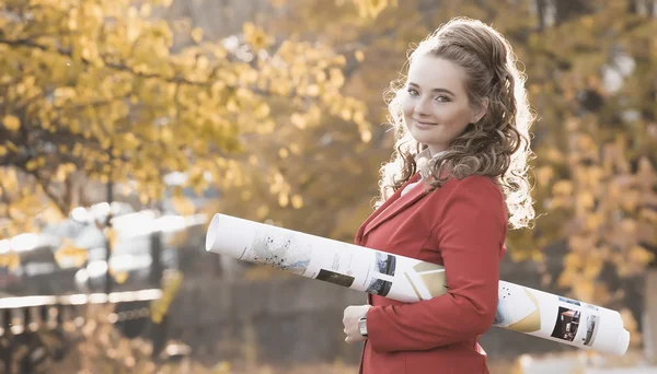 Young woman architect in red jacket and white helmet comes in the autumn on the street and keeps convolution with the drawings. backlight