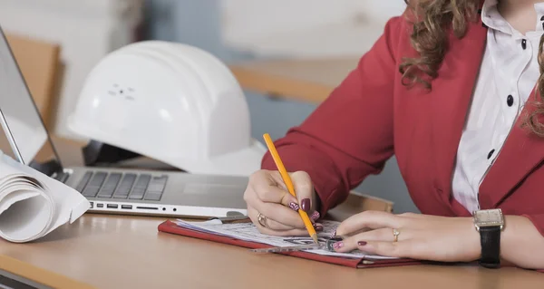 Young woman architect in red suit sitting at a table in front of the drawings. Computer is on the table. Female student with a pencil. Director.