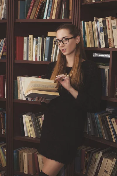 Young beautiful girl in a black dress standing in the library with books