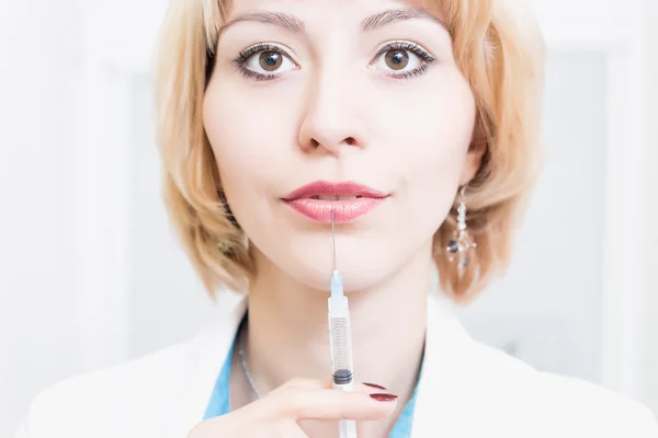 Woman beautician is permanent in a white robe in the office and keeps the needle of the syringe near the lips. Doctor with syringe