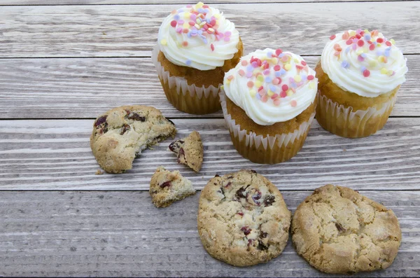 Tasty cookies and muffins cakes with vanilla cream decorated with sugar candies on a wooden background