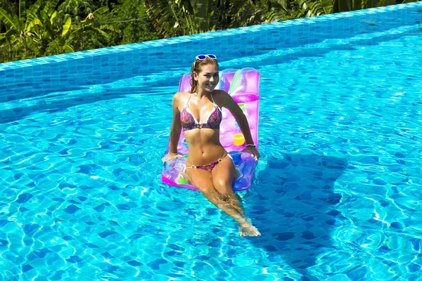 Woman wearing sunglasses and bikini poses swimming on mattress in pool. Phuket island, Thailand