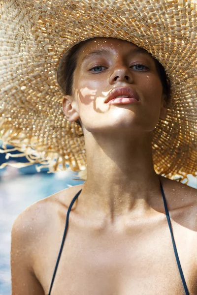Woman in hat and black stylish swimsuit poses near swimming pool enjoying sun. Phuket island, Thailand. High fashion look.