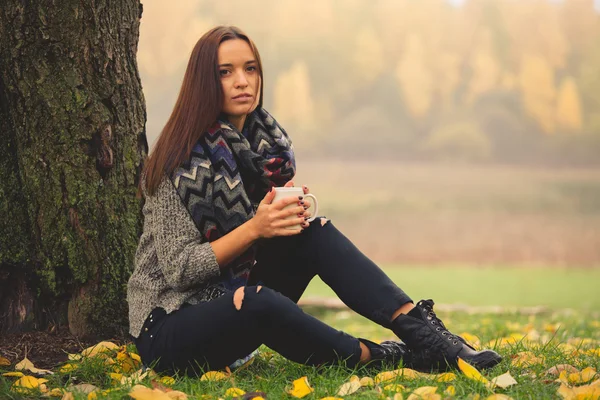 Beautiful woman having rest under tree with cup of tea. Lonely woman enjoying nature landscape in autumn. Autumn day. Girl sitting on grass Color horizontal image.