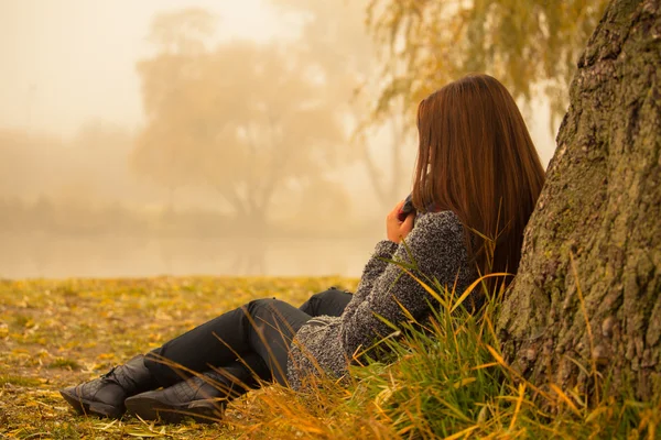 Lonely woman having rest under the tree near the water in a foggy autumn day. Lonely woman enjoying nature landscape in autumn. Autumn day. Girl sitting on grass horizontal image.