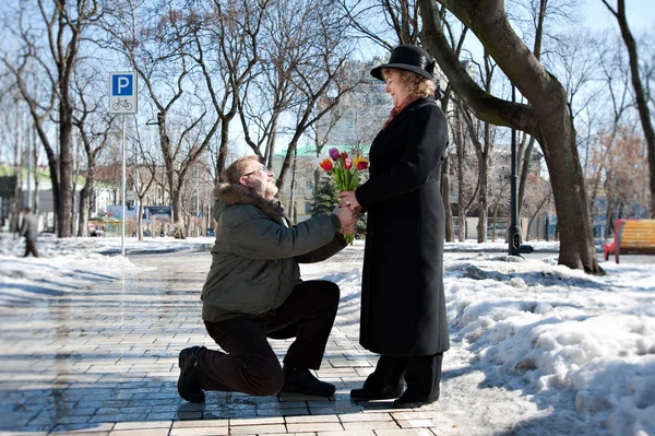 Old-aged man gives flowers to his woman, squatting on one leg