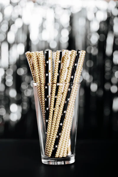 Striped mixed gold, black and white drink straws in glass on blurred background with black shiny bokeh
