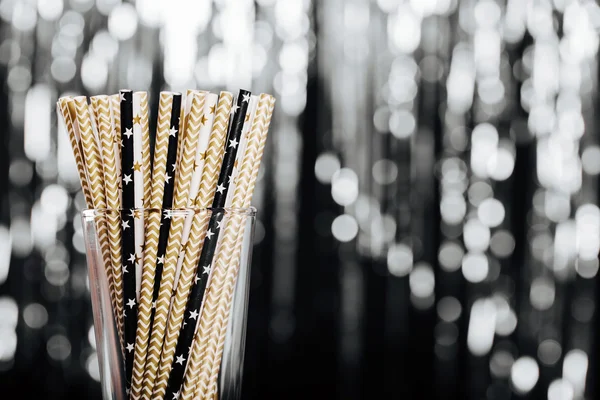 Striped mixed gold, black and white paper drink straws in glass on blurred background with black shiny bokeh