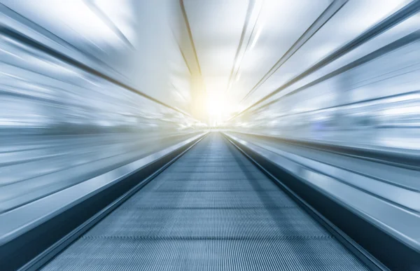 Perspective wide angle black and white view of modern light blue illuminated and spacious high-speed moving escalator with fast blurred trail of handrail in vanishing traffic motion
