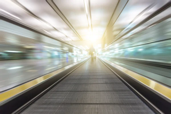 Perspective wide angle black and white view of modern light blue illuminated and spacious high-speed moving escalator with fast blurred trail of handrail in vanishing traffic motion