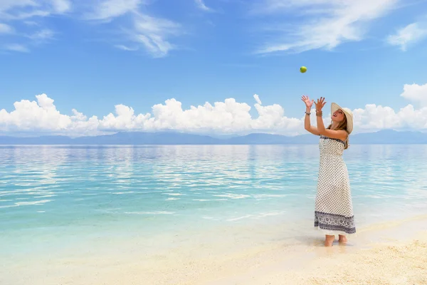 Youn europian woman in light dress and hat is walking on white sand beach near calm amazing sea playing with green apple at sunny day