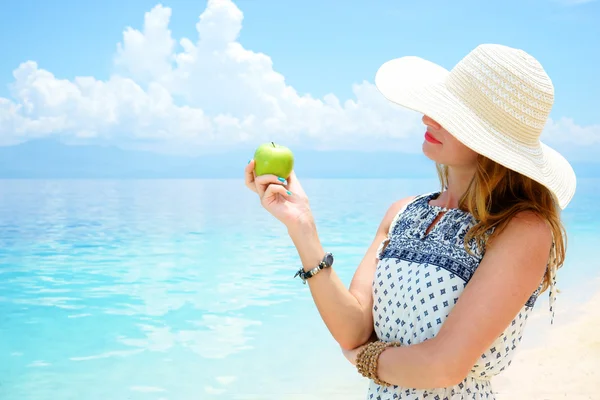 Young beautiful european woman is holding green apple in her hand against the calm soft tropical sea under tender blue sky