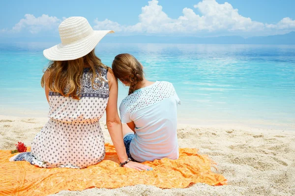 European mom with daughter are sitting together on white sand of tropical beach and looking at the turquoise sea under gentle cloudy sky