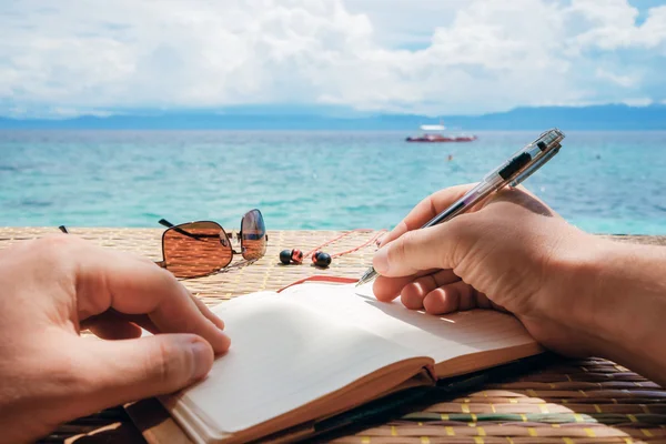 Caucasian man is writing sime idea, message or letter in his notepad by pen while he sitting on the beach of tropical sea with boat