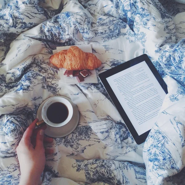 Cup of coffee in hand, eBook and croissant on bed