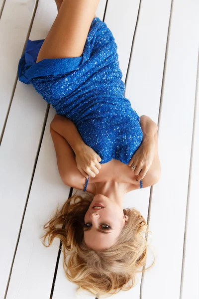 Sexy and beautiful woman in the blue evening sparkling dress with sequins is lying on the white wooden floor