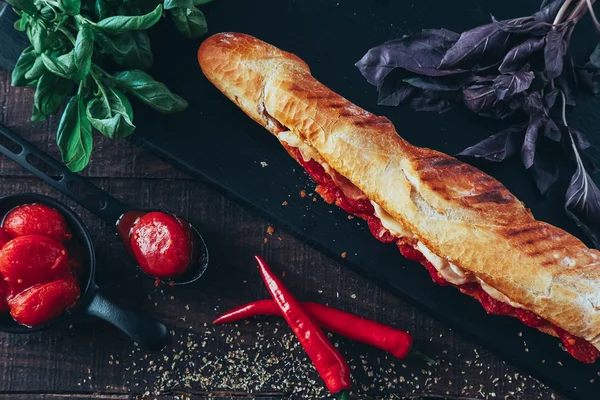 Long baguette sandwich with lettuce, vegetables, salami, chili and cheese on black background